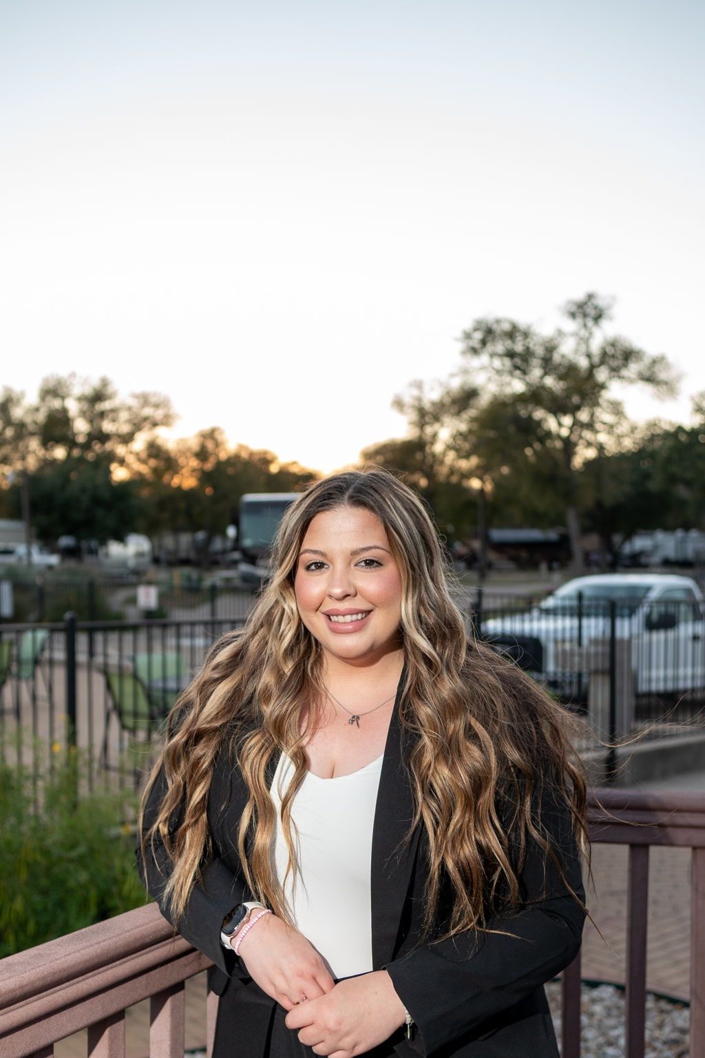 A woman with long wavy hair, wearing a black blazer and white top, stands outdoors by a wooden railing with trees and parked cars in the background at sunset.