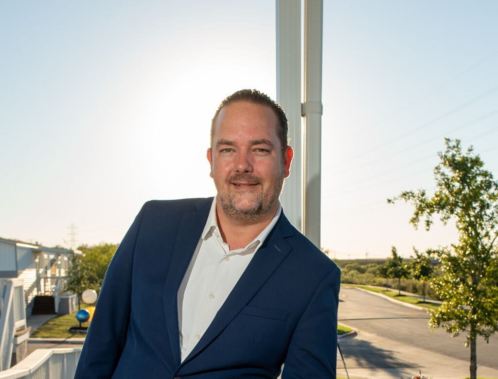 A man in a blue suit stands on a wooden porch, leaning on a white railing, with sunlight behind him and a suburban street in the background.
