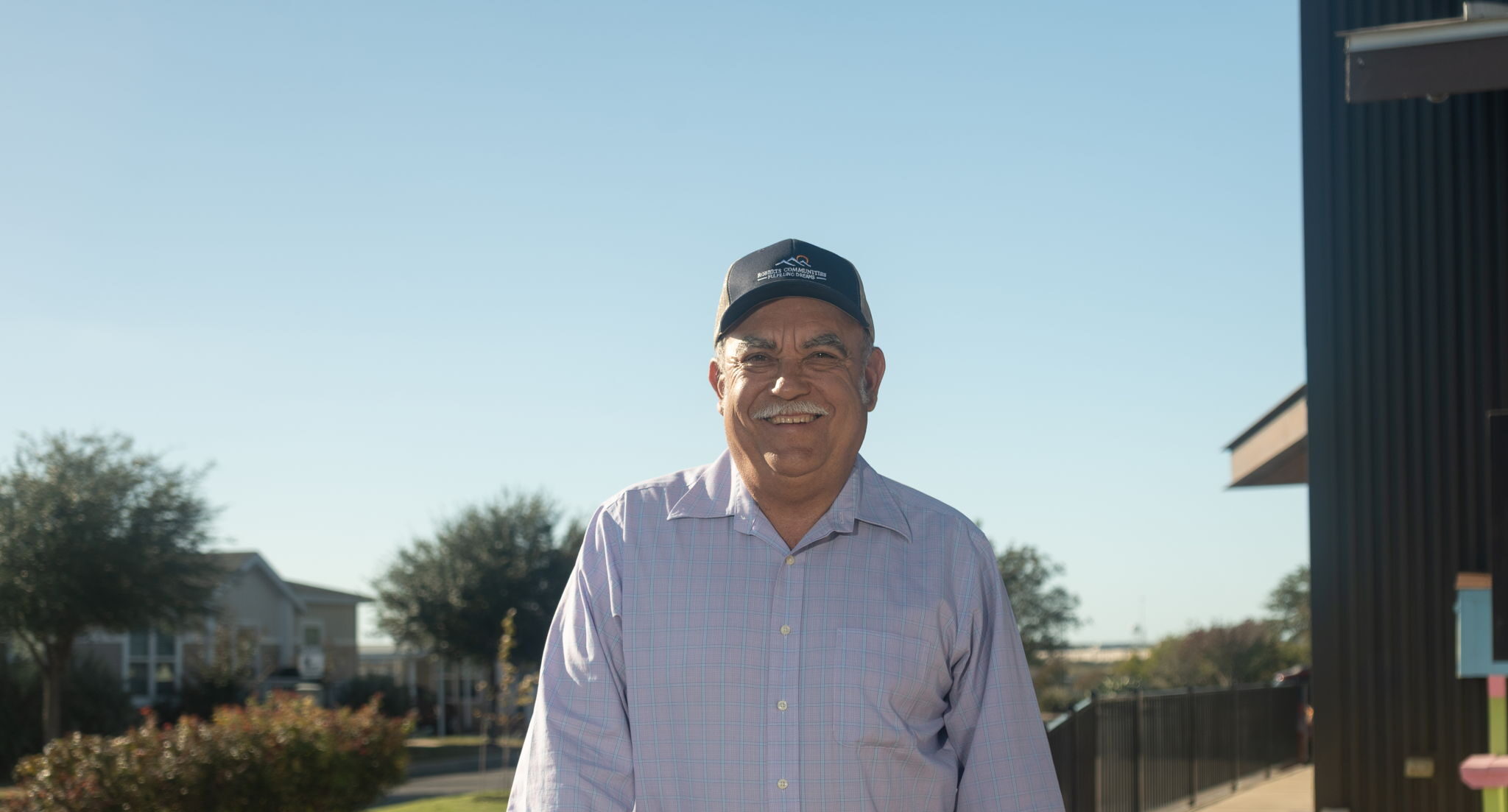 A man wearing a black cap and light purple shirt stands outdoors on a sunny day near a building and landscaped area.