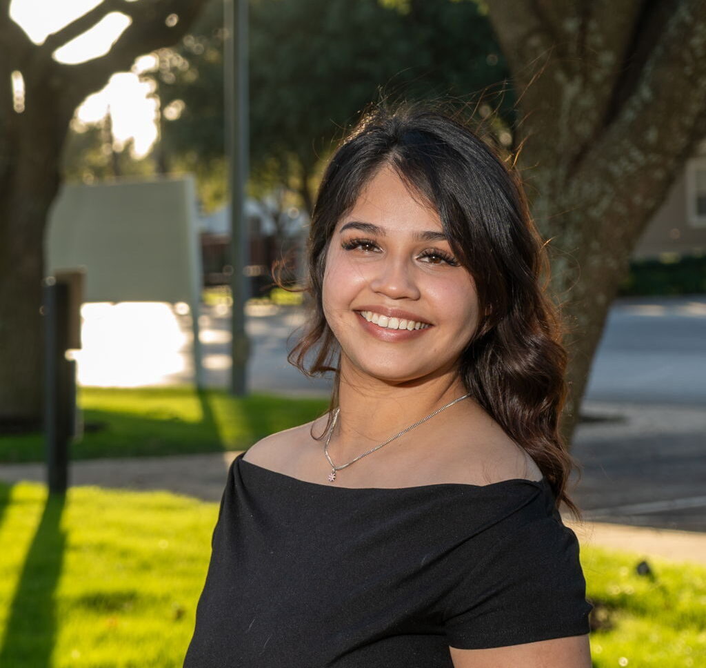 A woman in a black off-shoulder top stands outside on a sunny day, smiling, with trees and grass in the background.