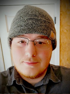 A person with glasses and a short beard wears a gray knit beanie and a dark collared shirt, standing indoors in front of a white door and a wood-paneled wall.