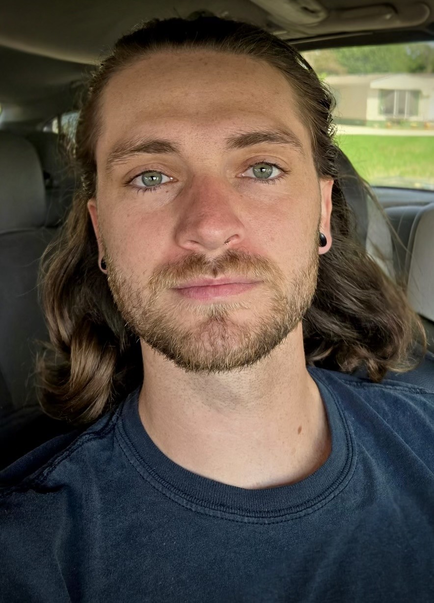A man with long brown hair, a beard, and blue eyes sits in a car, wearing a dark T-shirt, looking directly at the camera.