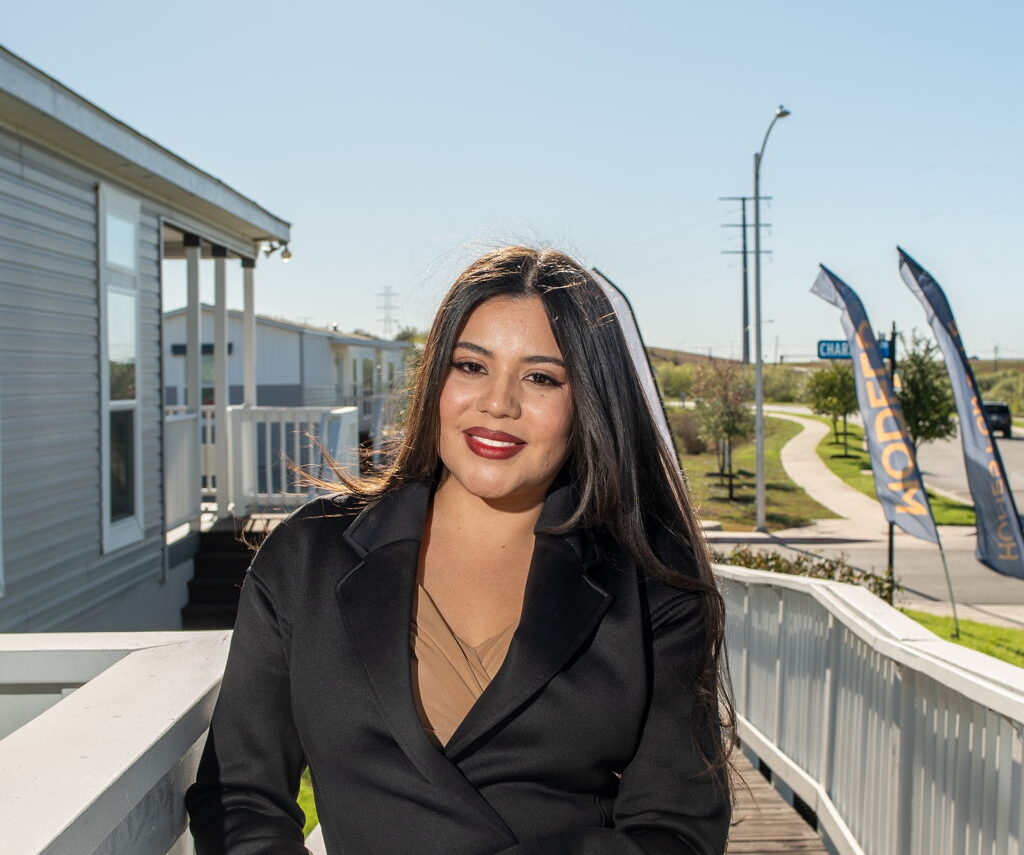 A woman in a black blazer stands on a wooden walkway outside a building, smiling at the camera on a sunny day.