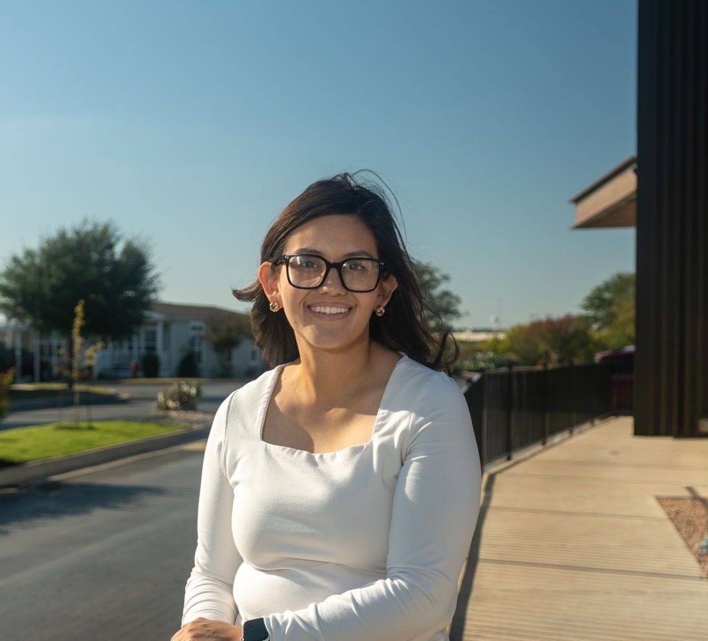 A woman wearing glasses and a white long-sleeve shirt stands outside, smiling, with a clear blue sky and buildings in the background.
