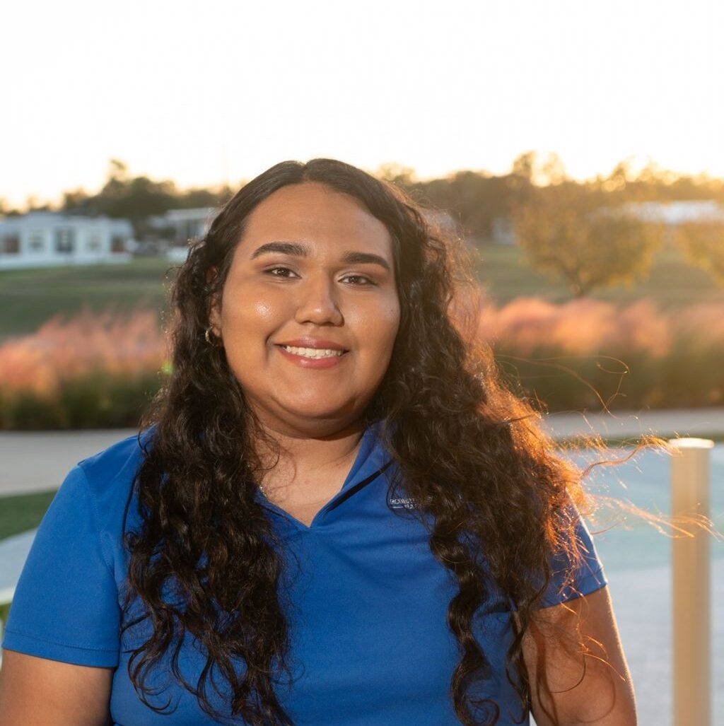 A person with long curly hair, wearing a blue polo shirt, smiles outdoors with sunlight and greenery in the background.