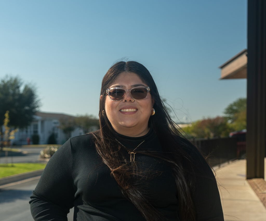 A woman with long dark hair, sunglasses, and a black outfit stands outdoors near a building, smiling at the camera on a sunny day.