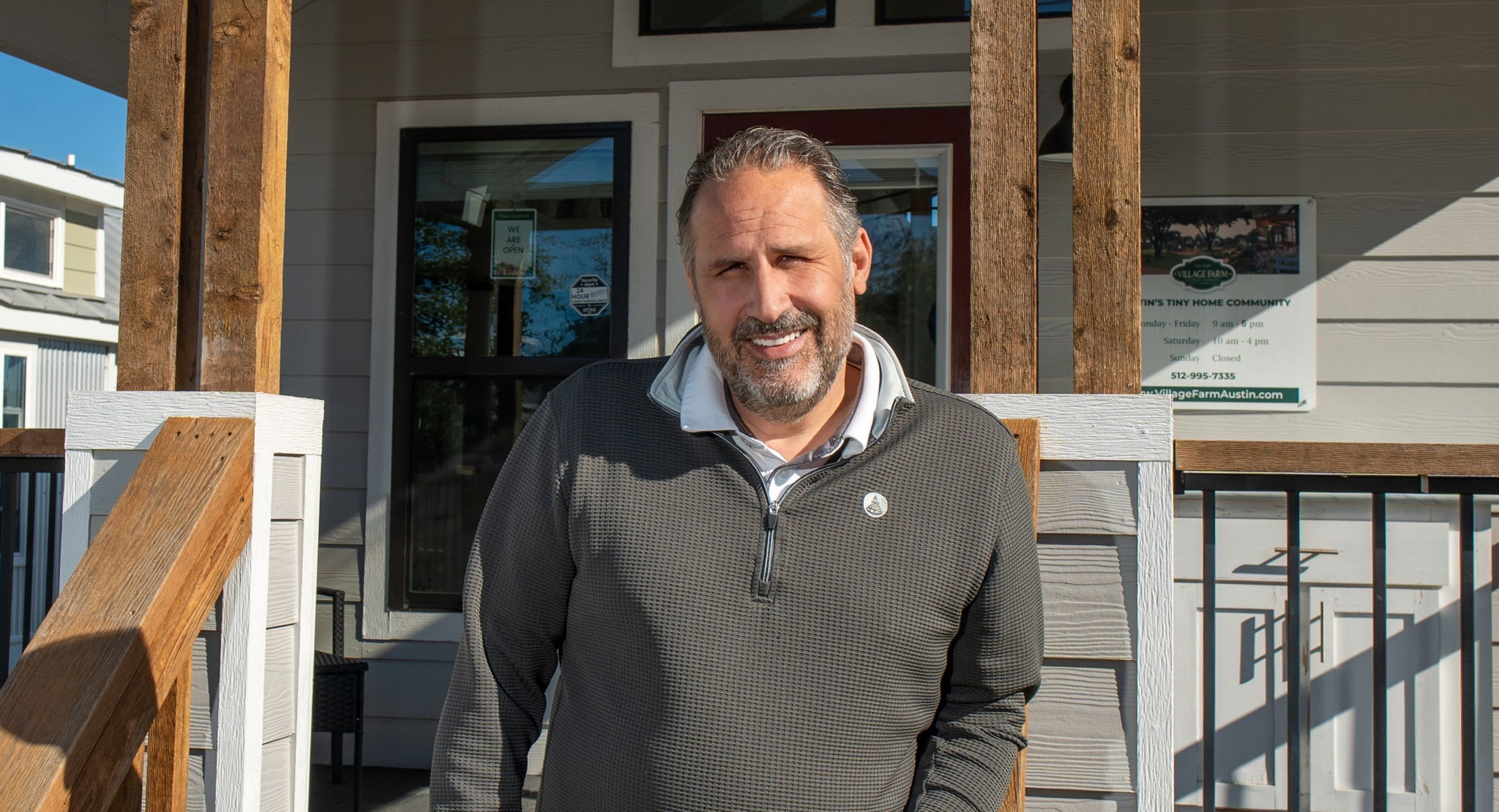 A man stands on the porch of a modern house, leaning against the wooden railing, with signage visible in the background.