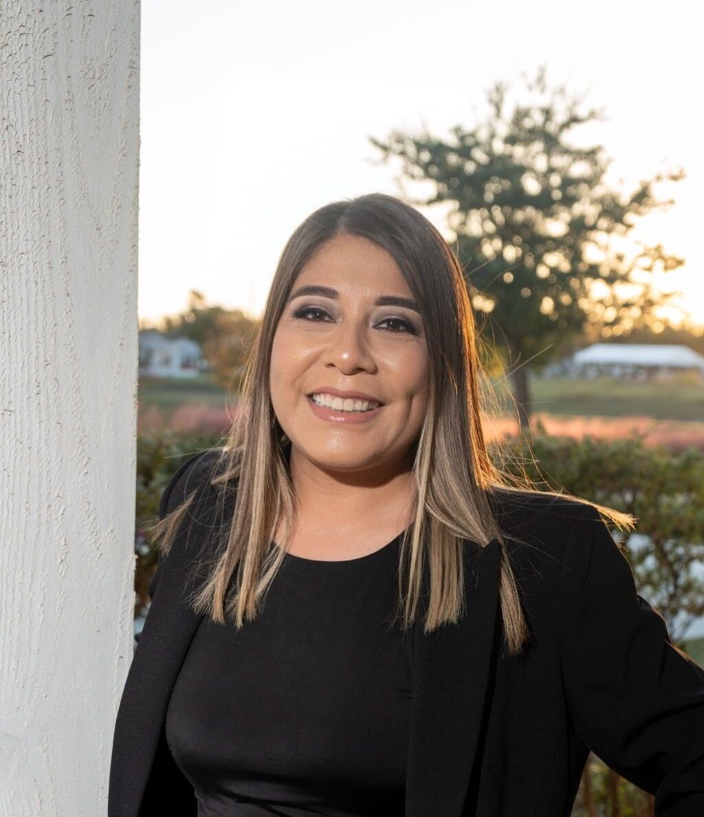 A woman with straight, light brown hair is smiling, wearing a black blazer and top with gray pants, standing outdoors near a white pillar at sunset.
