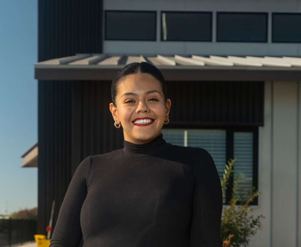 A woman wearing a black long-sleeve top stands and smiles in front of a modern building under a clear blue sky.