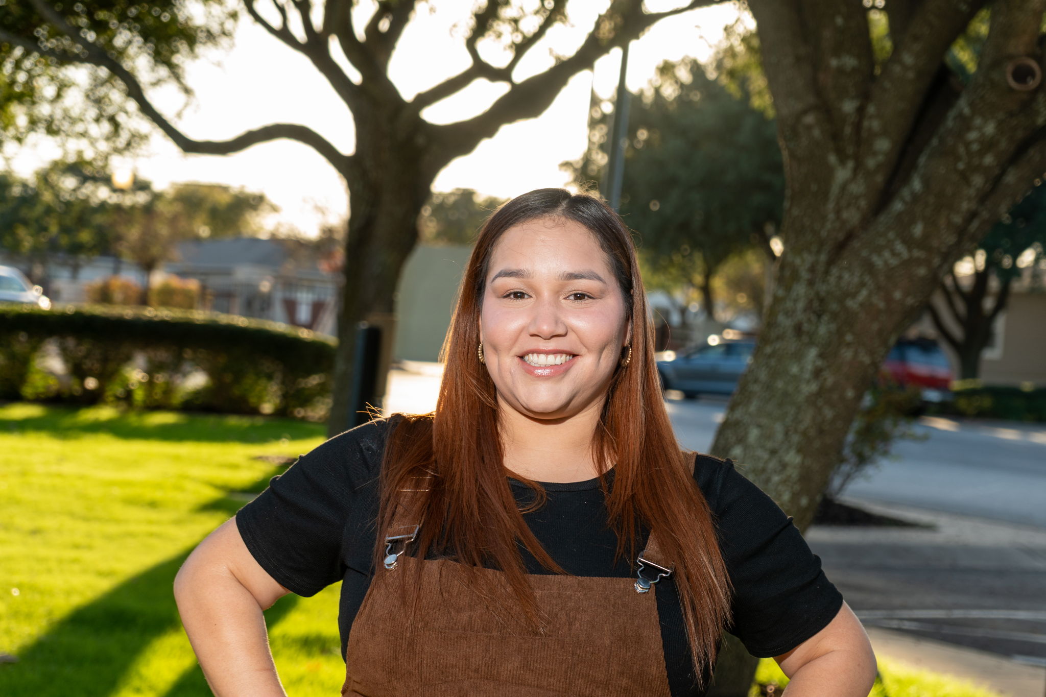 A woman with long brown hair stands outdoors, smiling at the camera, with trees, grass, and a street in the background.