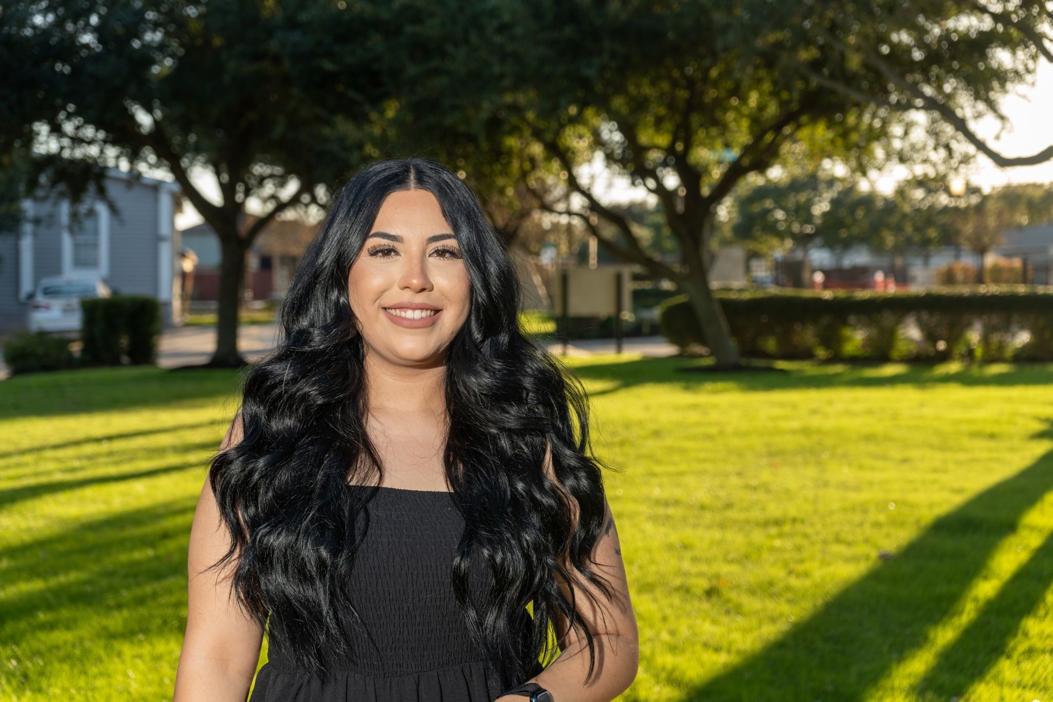 A woman with long black hair, wearing a black dress, stands smiling on a green lawn with trees and sunlight in the background.