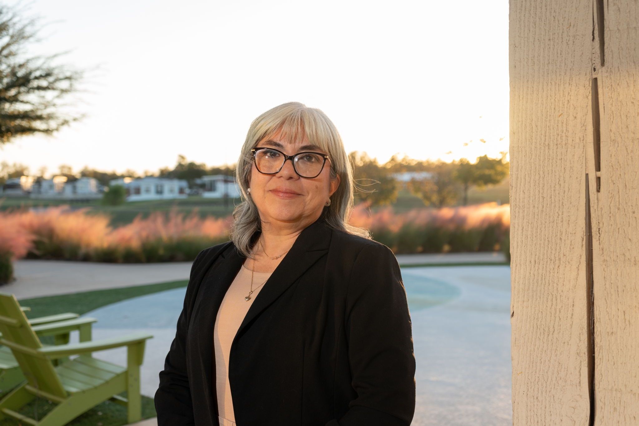 A woman with gray hair and glasses stands outdoors near a wooden post, wearing a black blazer, with a lawn, chairs, and trees in the background at sunset.