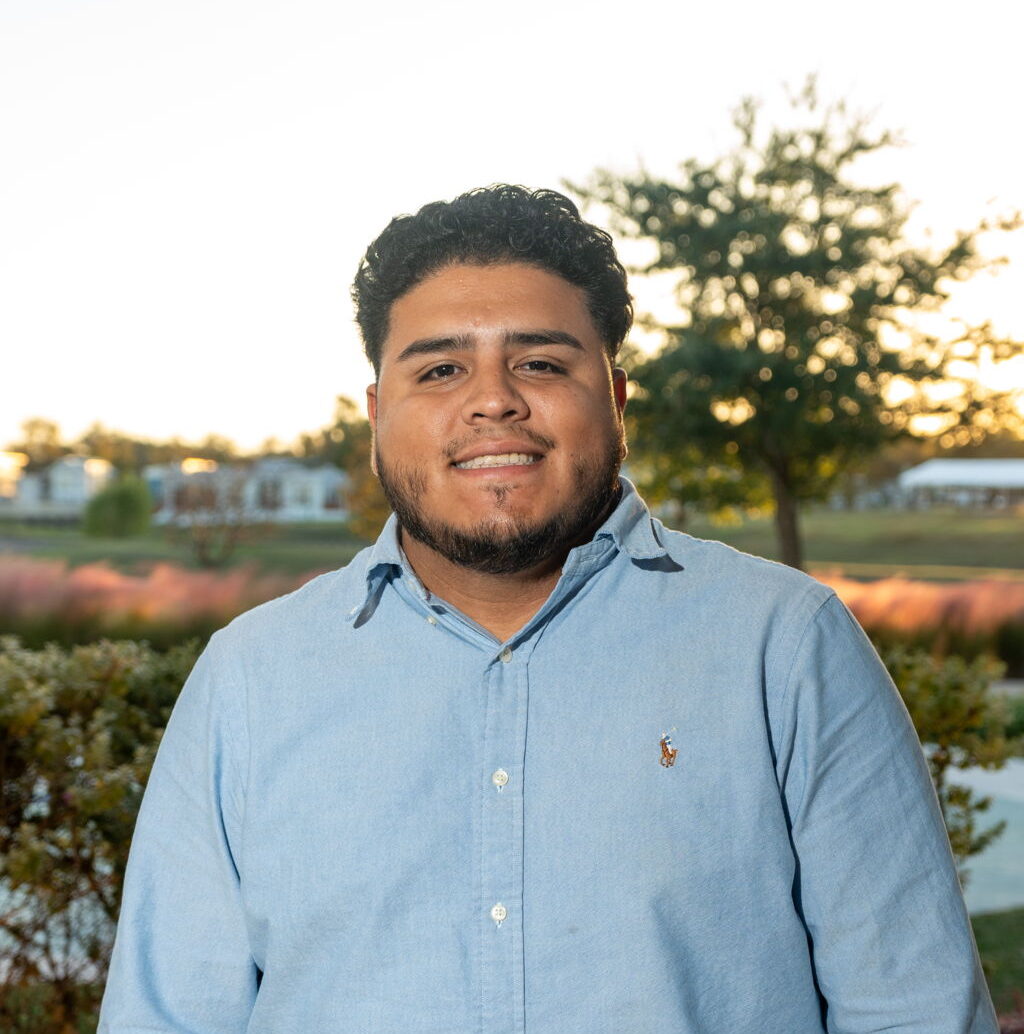 A man with short curly hair and a beard, wearing a light blue button-up shirt, stands outdoors in front of bushes and trees at sunset.
