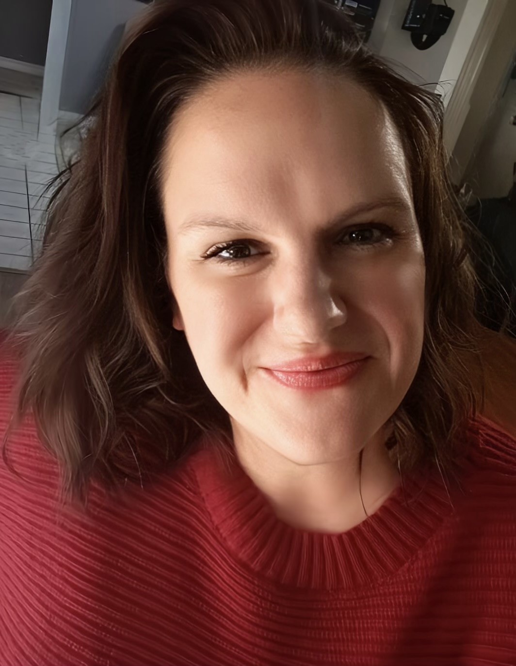 A woman with shoulder-length brown hair and a red sweater smiles at the camera indoors, with a tiled floor and hallway visible in the background.