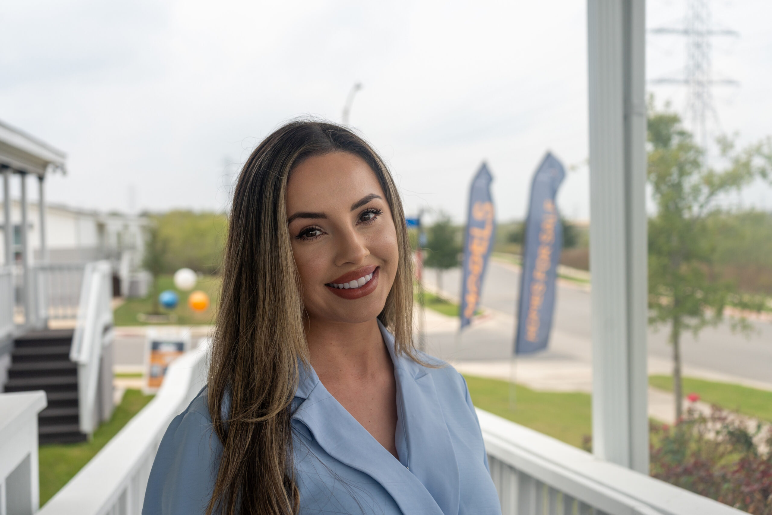 A woman with long brown hair and a blue blouse smiles while standing on a porch with a street and banners visible in the background.