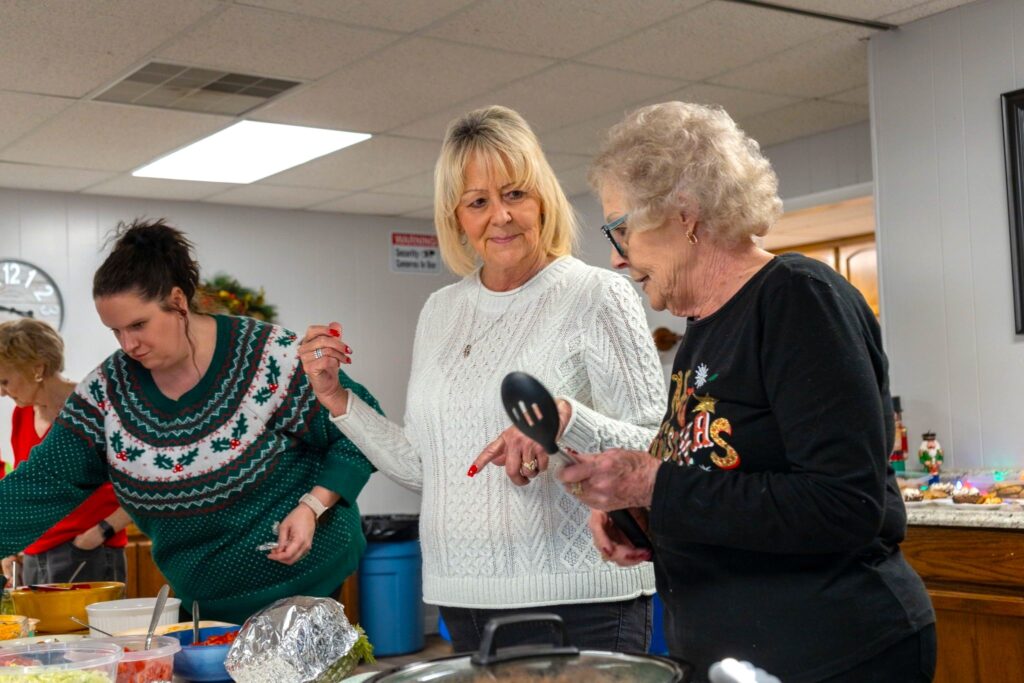 Three women stand at a table with various dishes, serving food in a brightly lit room decorated for the holidays.