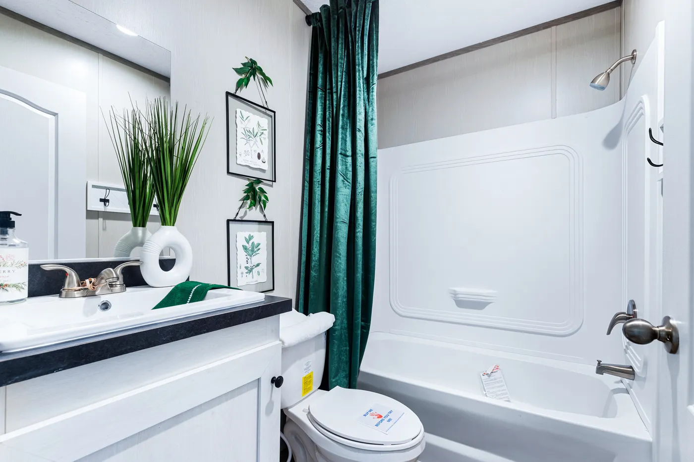 Modern bathroom with a white vanity, potted plant, framed botanical art, green shower curtain, and white bathtub with shower.