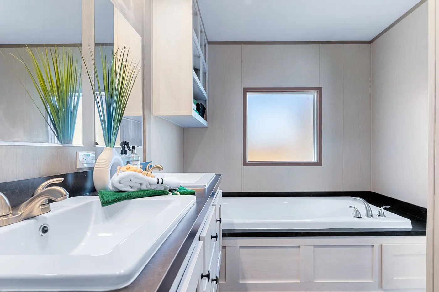 Modern bathroom with a white sink, gold faucet, large mirror, built-in shelves, decorative plants, and a rectangular bathtub next to a frosted window.