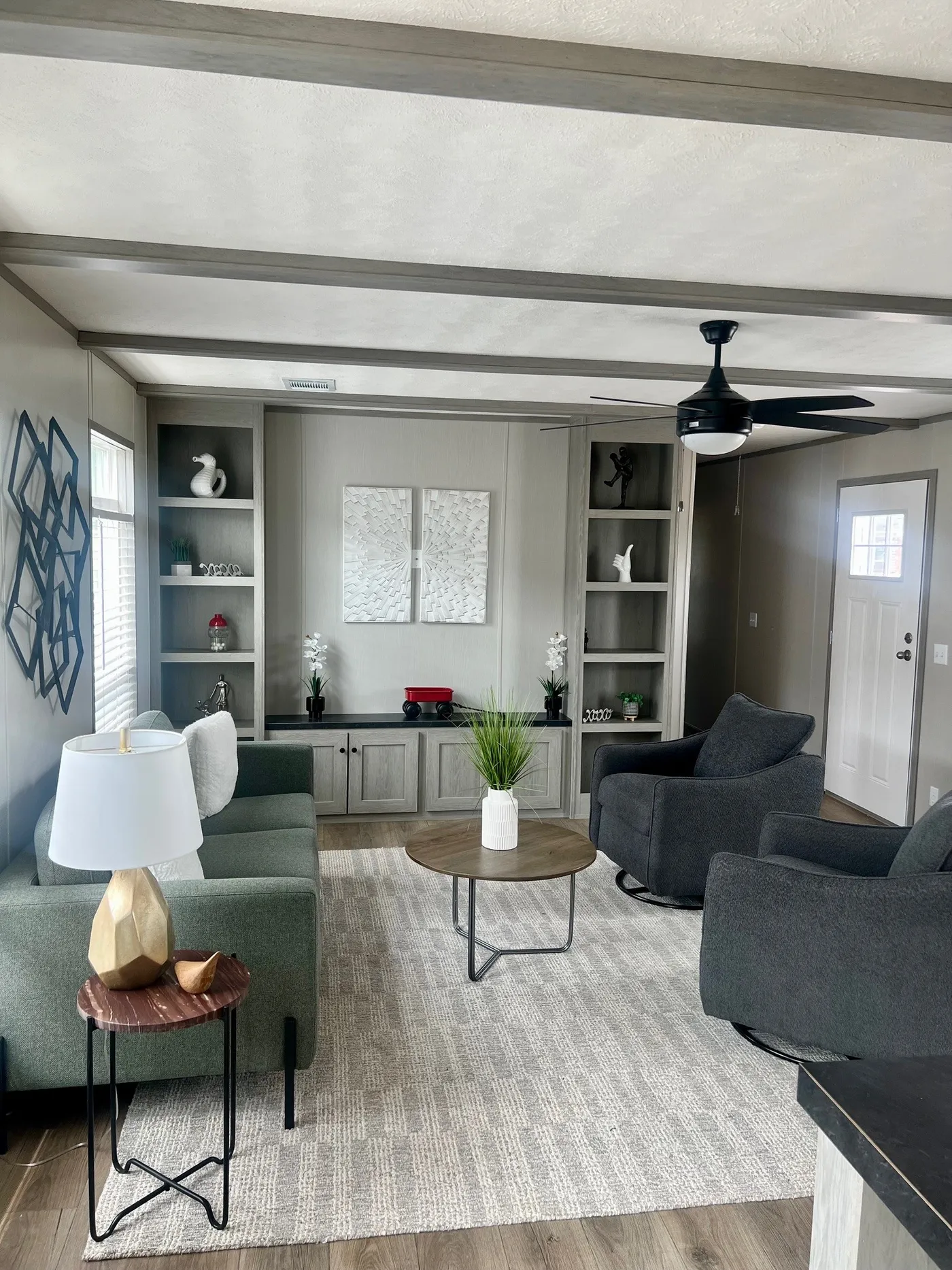 Modern living room with gray and green furniture, a round coffee table, built-in shelves, wall art, neutral decor, and a ceiling fan, viewed from one corner of the space.