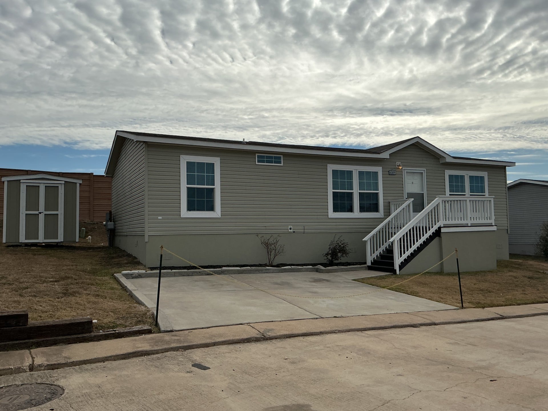 A beige manufactured home with white trim and steps, a small shed to the left, and a concrete driveway under a partly cloudy sky.