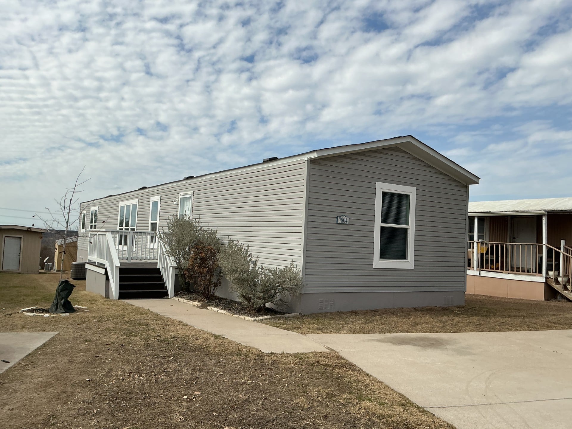 A light gray manufactured home with a ramp and steps leading to the entrance, set on a grassy lot with a concrete walkway under a partly cloudy sky.