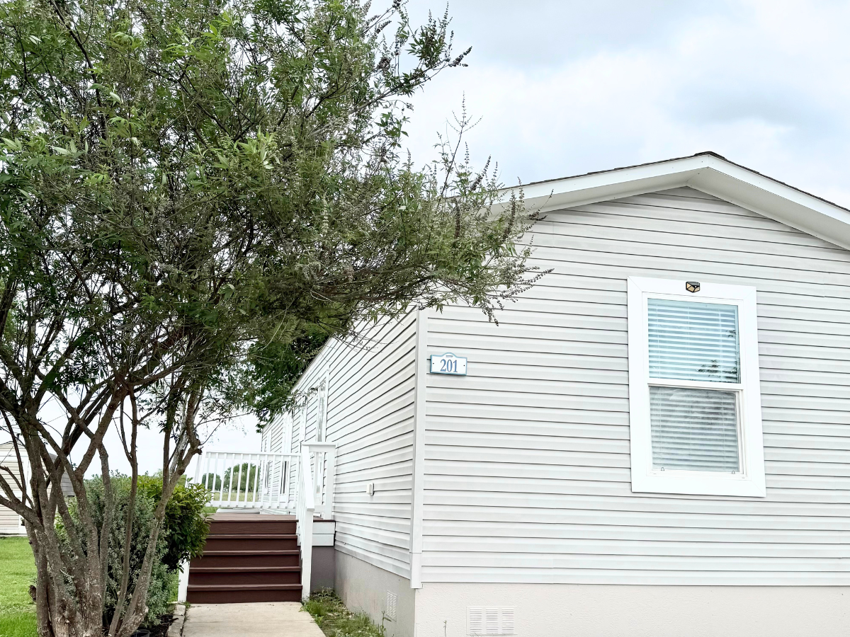 A white manufactured home with a front porch, stairs, and the house number 201. A tree is in the foreground next to the walkway.