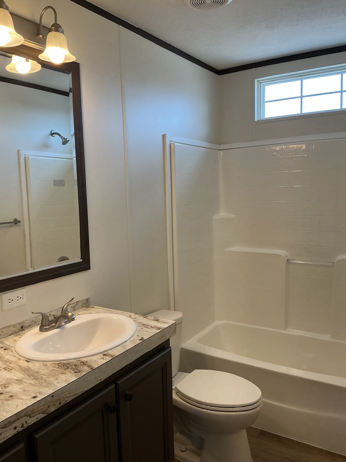 A bathroom with a sink, marble countertop, mirror with lights, toilet, and a white bathtub-shower combo, with a small window above the shower.