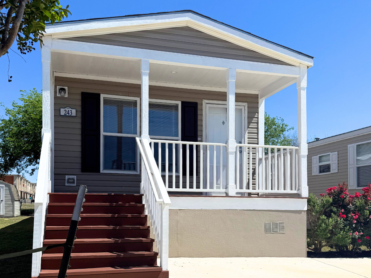 A small, single-story manufactured home with a front porch, white railing, and red steps, located in a residential area with clear blue sky.