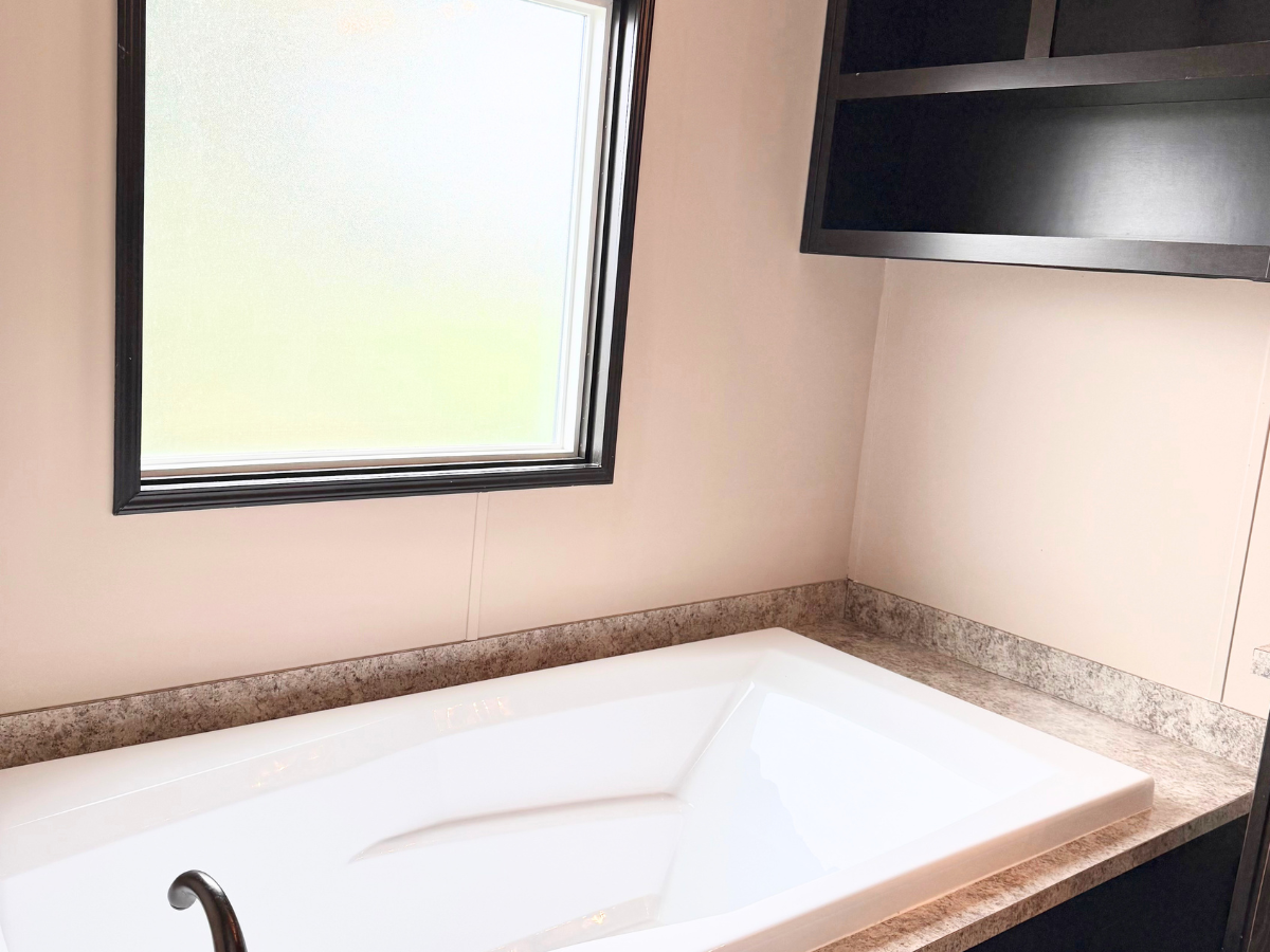 A white bathtub with a dark faucet is set against a beige wall under a frosted window, with dark shelves mounted above the tub.