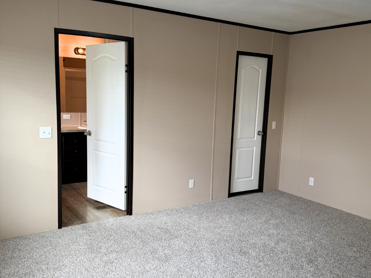 Empty beige room with carpet, two white doors—one open showing a bathroom vanity, the other closed. Walls and baseboards have a dark trim.