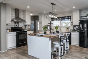 Modern kitchen with white cabinets, black appliances, a large island with bar stools, wooden countertops, and contemporary lighting fixtures. Plants and decorative items are on the counters.