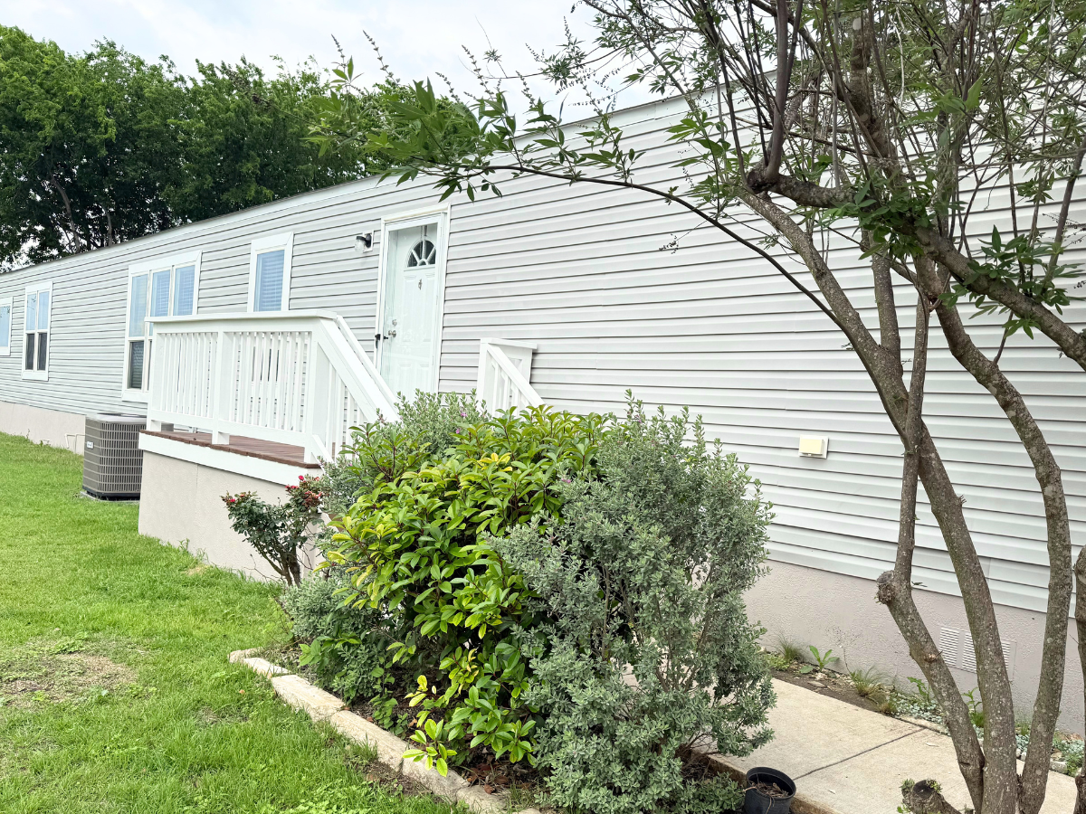 A white mobile home with a small porch and steps is surrounded by green shrubs and a tree on a grassy lawn.