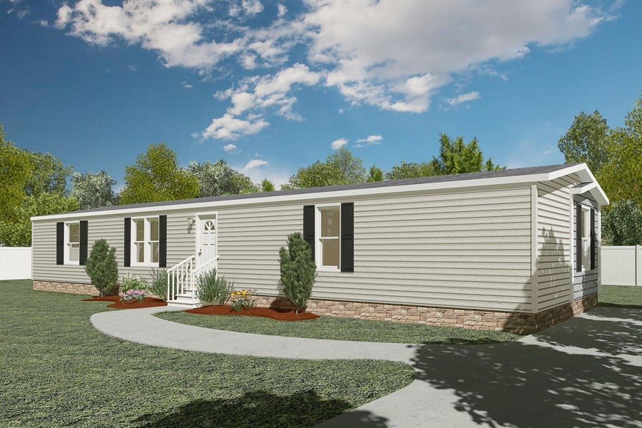 Single-story manufactured home with gray siding, black shutters, and a small landscaped front yard with shrubs, against a backdrop of trees and a blue sky.