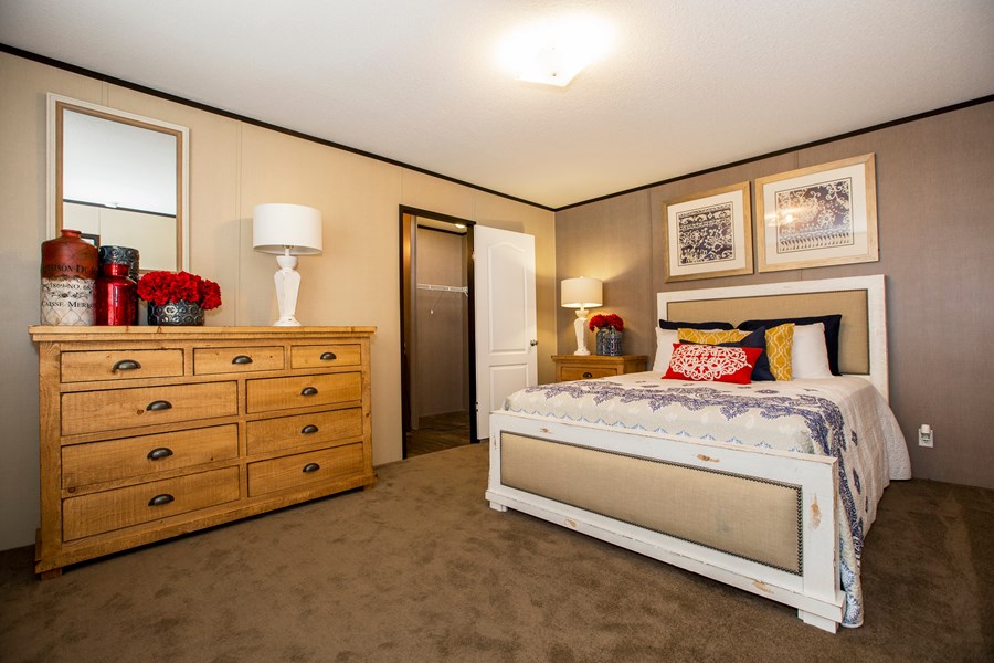 A bedroom with a double bed, wooden dresser, wall art above the bed, two mirrors, and decorative vases and flowers on the dresser. A closet with an open door is visible.