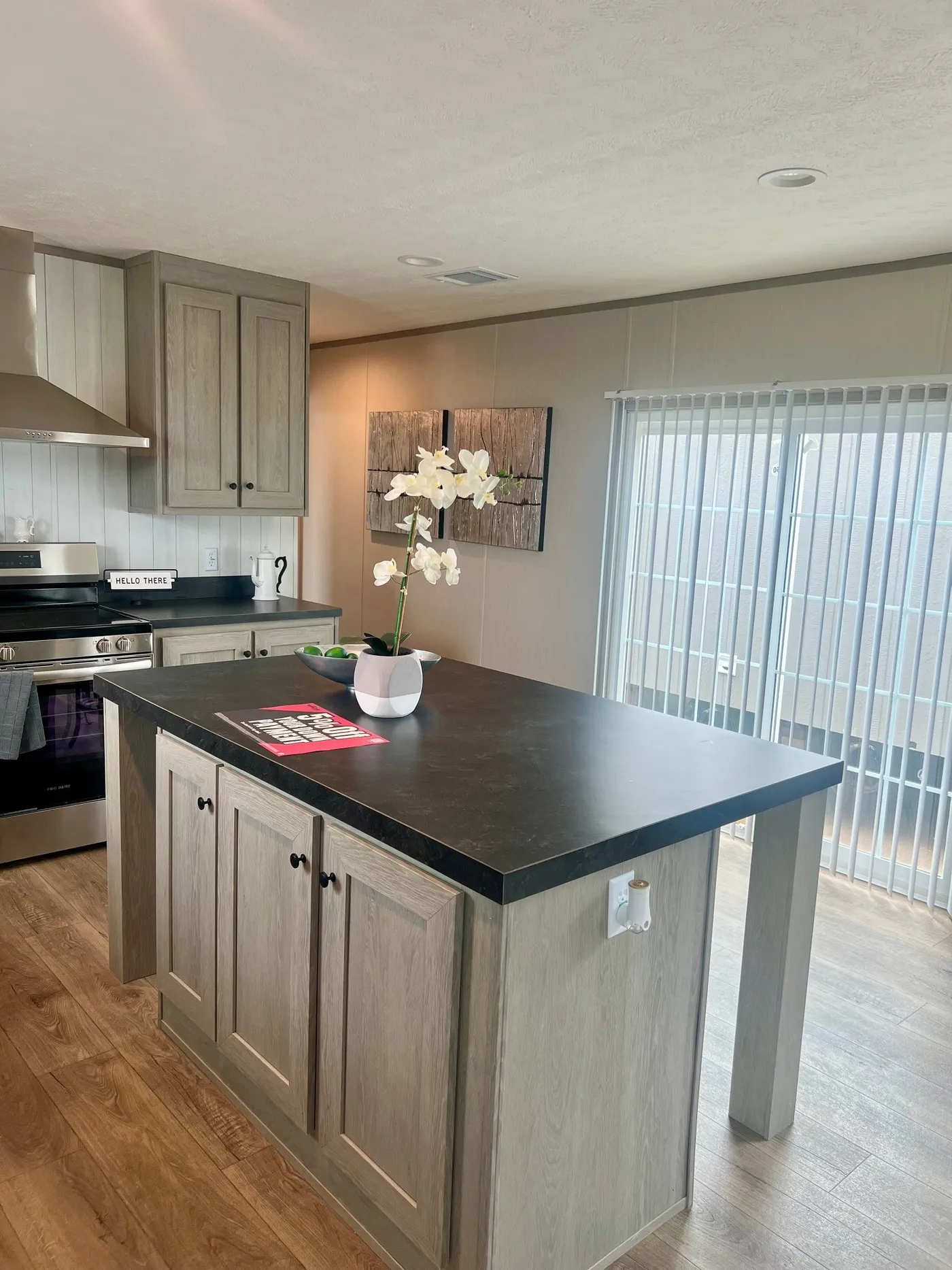 A modern kitchen with light wood cabinets, a black countertop island, a potted orchid, stainless steel appliances, and vertical blinds covering a sliding glass door.