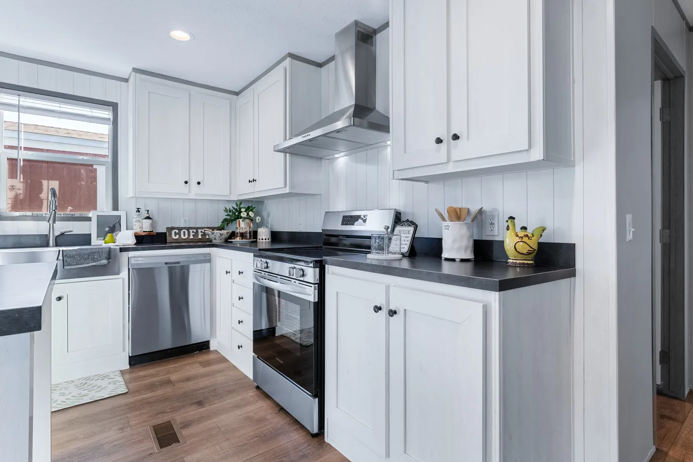 Modern kitchen with white cabinets, stainless steel appliances, black countertops, and wooden flooring. A yellow teapot sits near the stove.