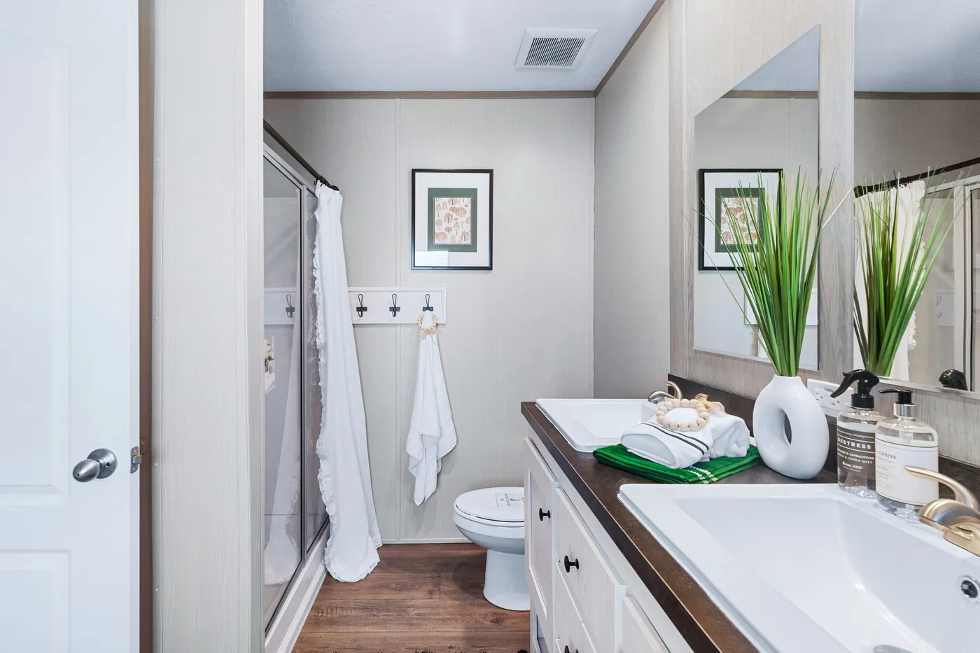 A bathroom with a double sink vanity, a toilet, a glass shower with a white curtain, wall hooks with towels, and framed artwork on beige walls.