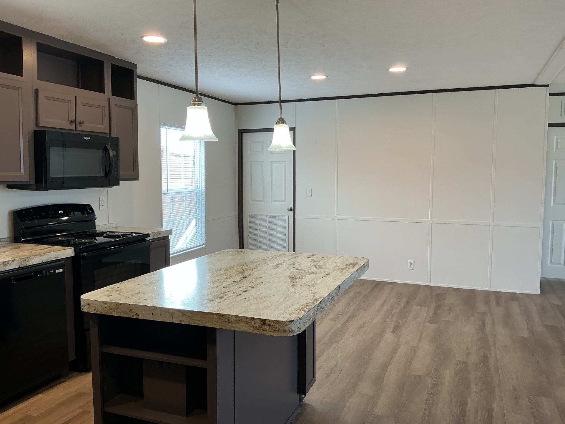 A modern kitchen with a marble island, black appliances, brown cabinets, wood flooring, pendant lights, and a door leading to an adjacent room.
