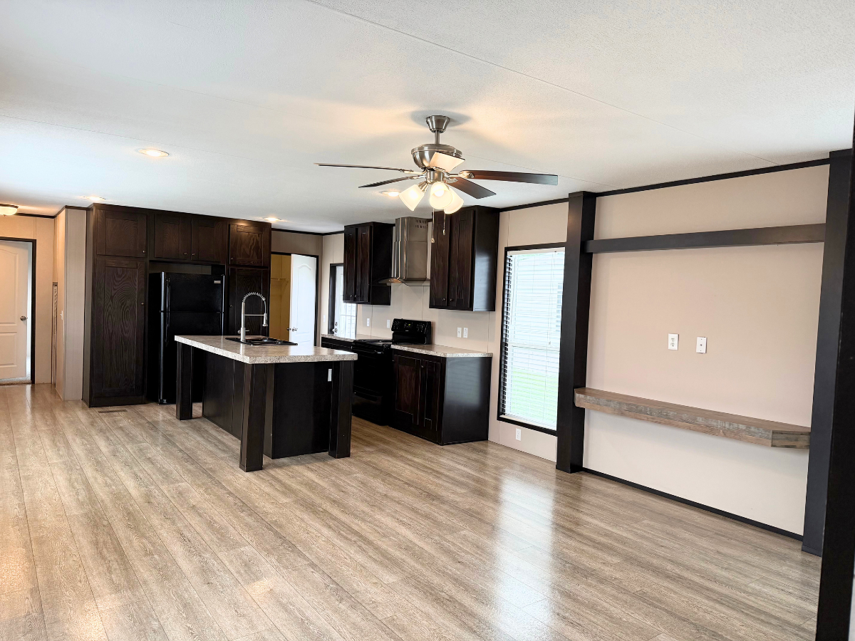 Modern open kitchen with dark cabinets, black appliances, an island with a sink, ceiling fan, and built-in shelving in a spacious room with light wood flooring.