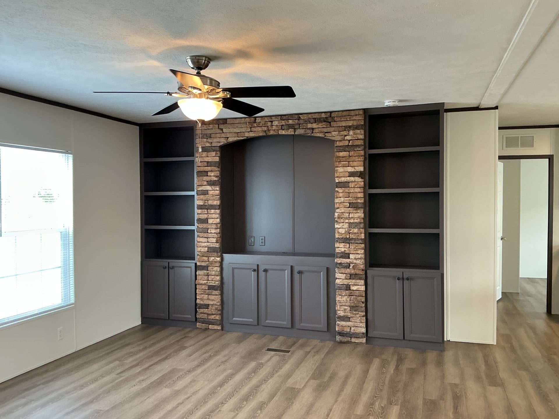 Built-in shelving and cabinets with a stone accent surround a recessed area in a modern living room with wood flooring, a ceiling fan, and large window.