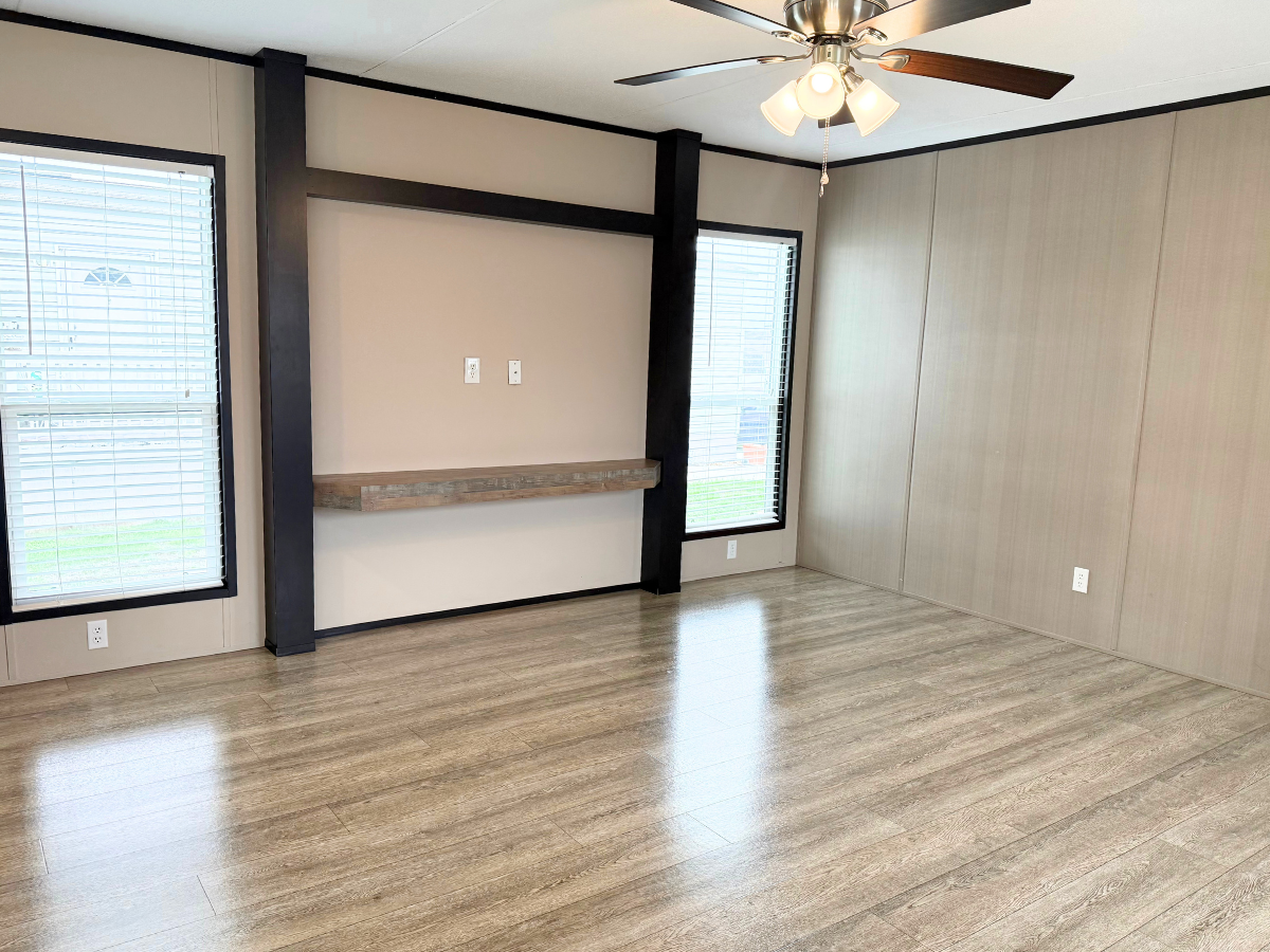 Empty room with light wood flooring, beige walls, two large windows with blinds, a ceiling fan with lights, and a wall shelf under electrical outlets.