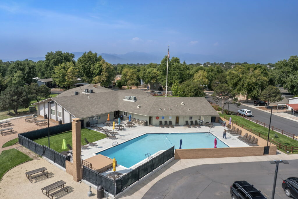 Aerial view of a community swimming pool with lounge chairs, shaded tables, and a single-story clubhouse surrounded by trees and parked cars.