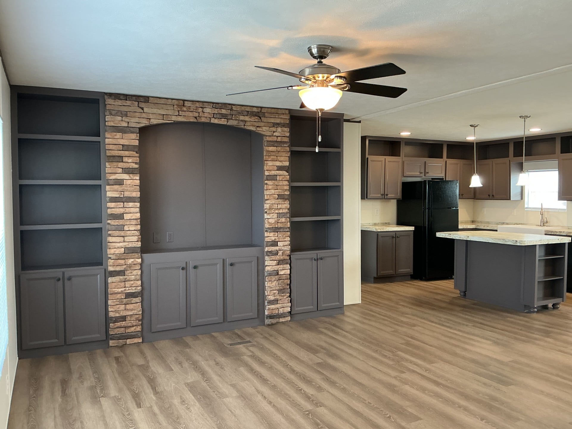 Open-concept living space with gray built-in shelves, stone accent wall, ceiling fan, wood-look flooring, and a kitchen with dark cabinets and an island.