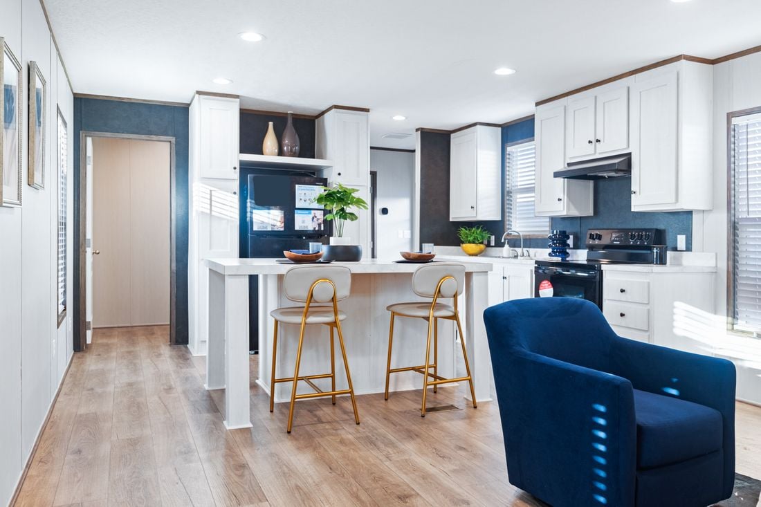 Bright modern kitchen with white cabinets, black appliances, a kitchen island with two gold stools, and a blue armchair on wood flooring.