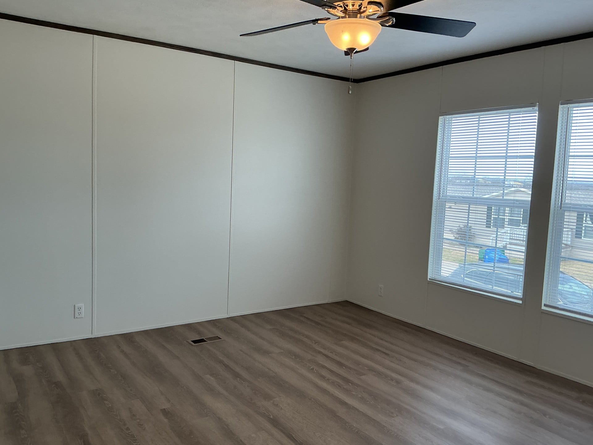 Empty room with light-colored walls, wood-look flooring, a ceiling fan with a light, and two large windows letting in natural light.
