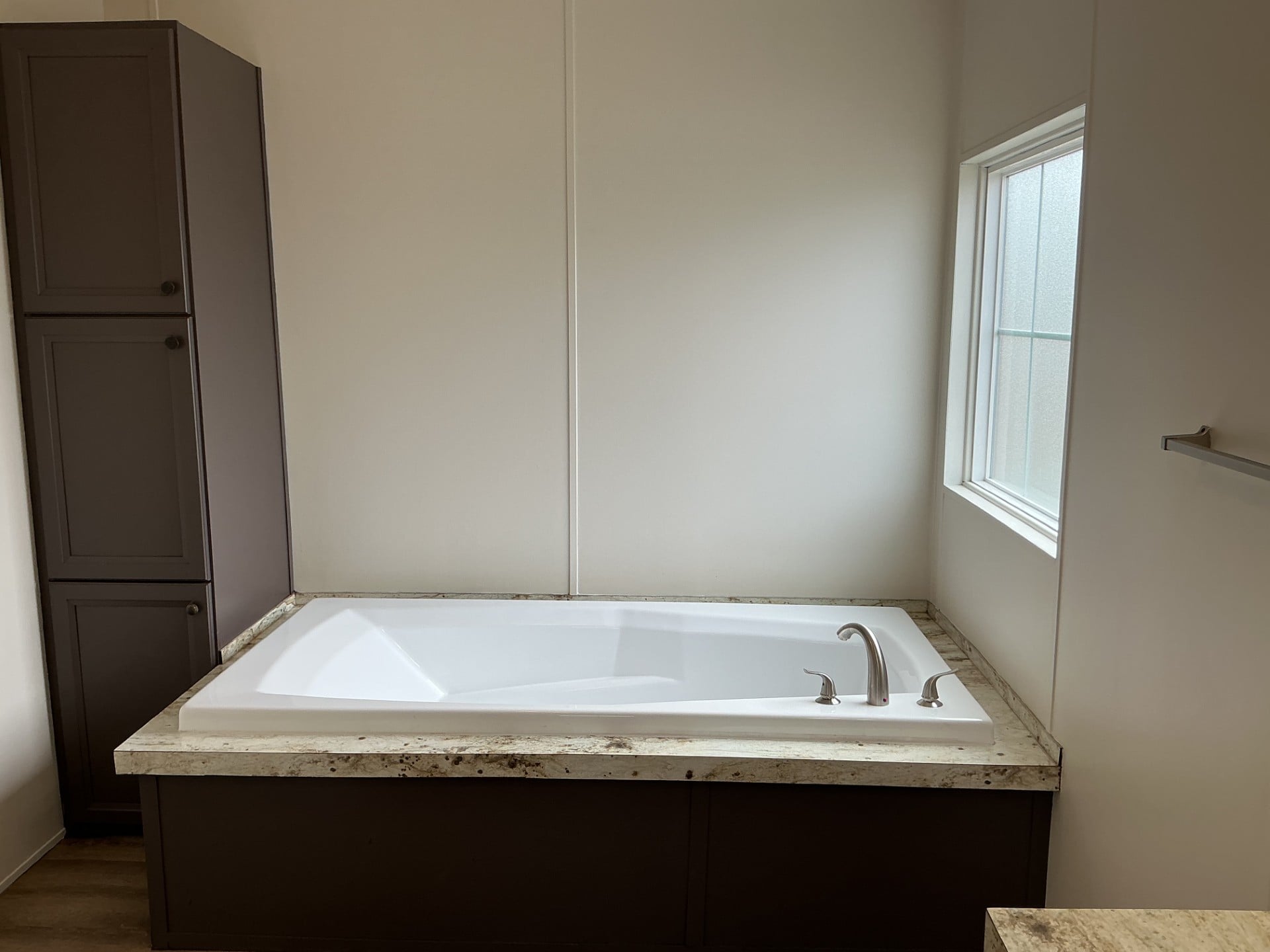 A built-in rectangular bathtub with marble trim is positioned near a window and next to a tall storage cabinet in a modern bathroom.