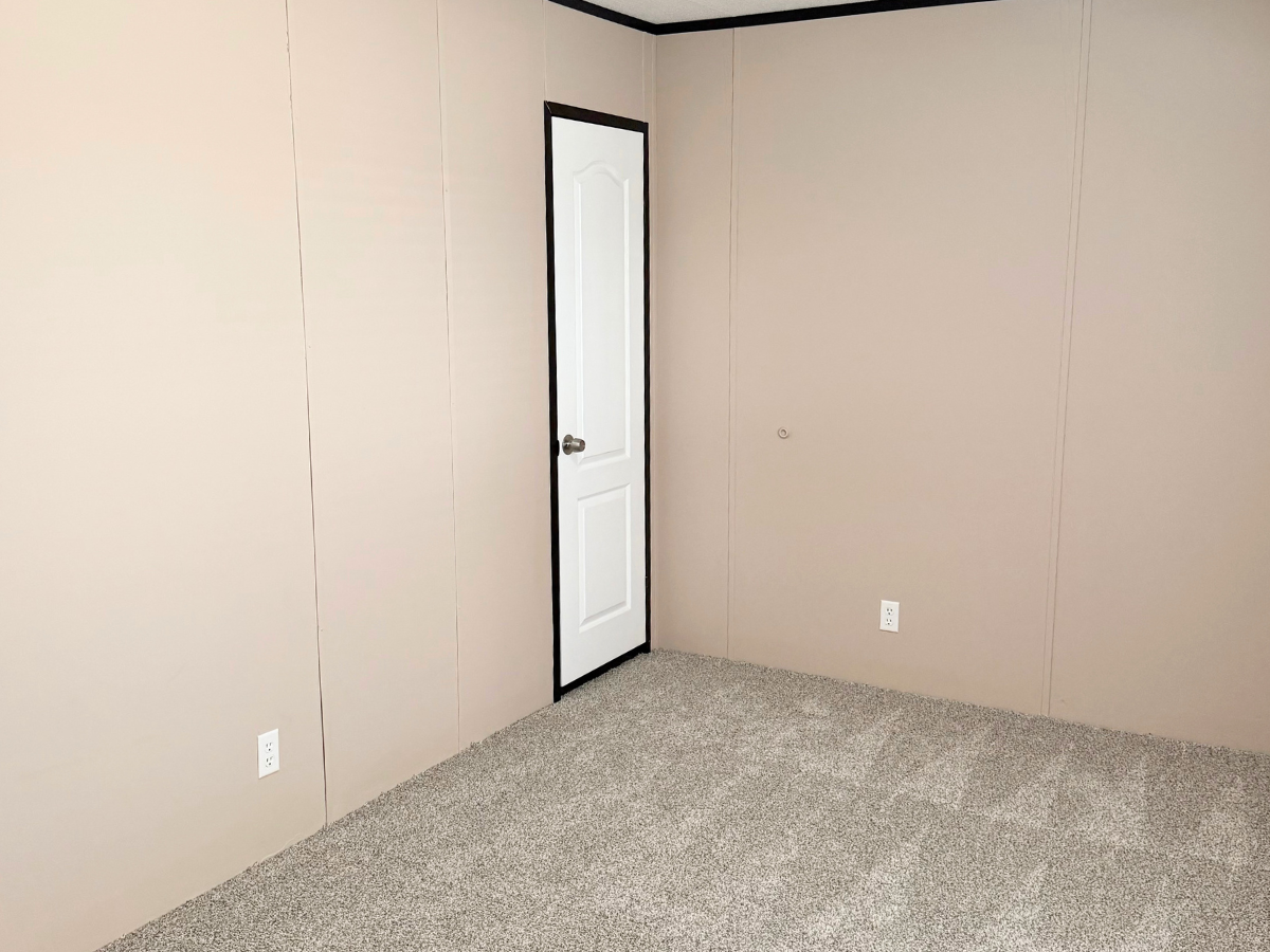 A small, empty room with beige walls, a white door, gray carpet, and two electrical outlets visible on the walls.