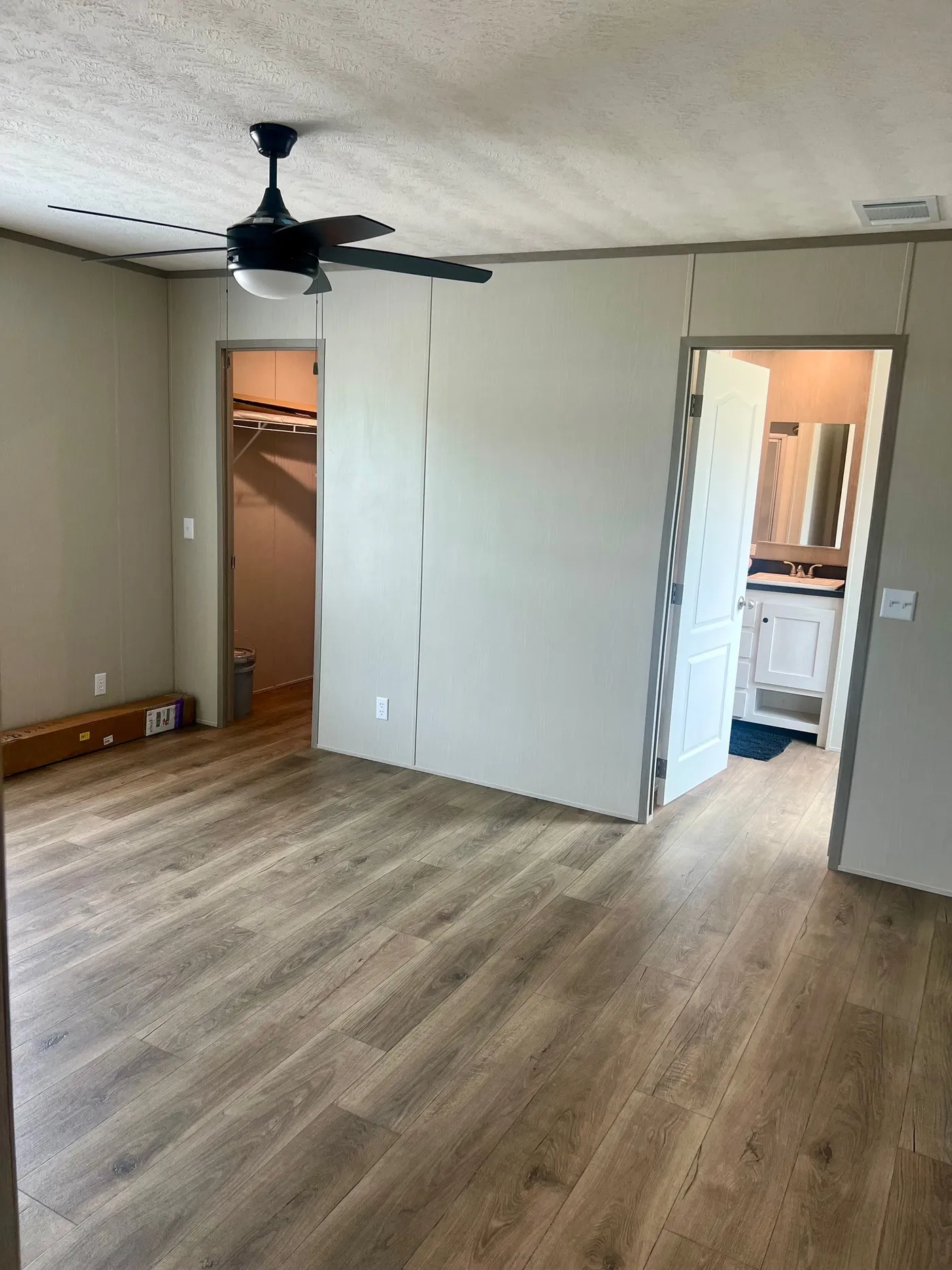Empty room with light wood flooring, ceiling fan, open closet, and open door leading to a bathroom with visible sink and mirror.