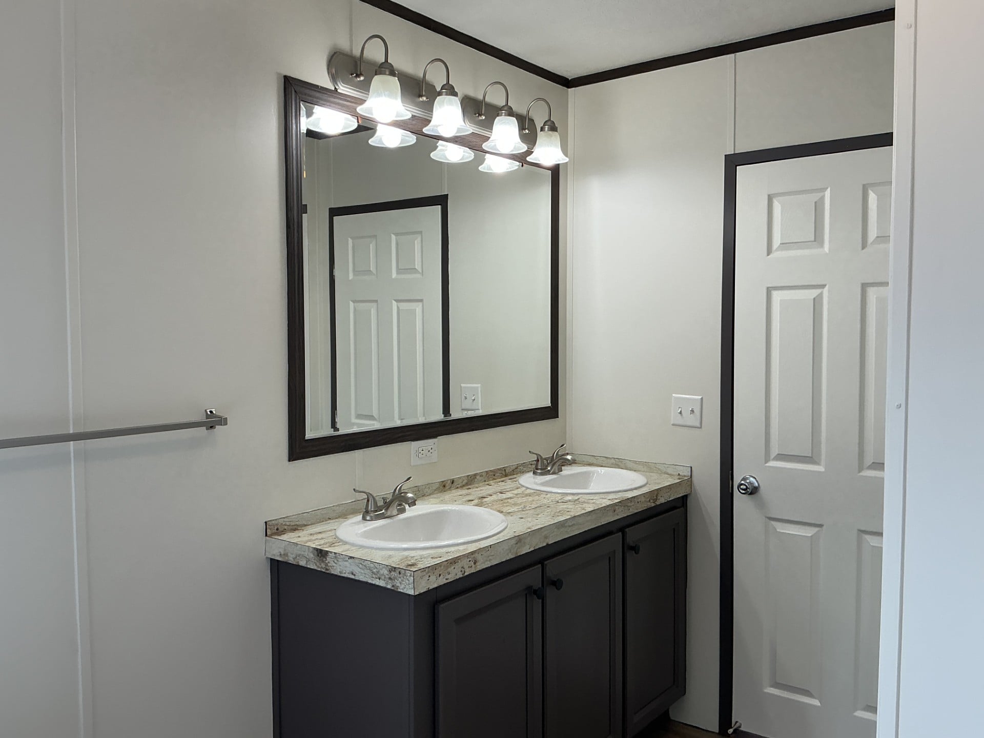 Double sink bathroom vanity with dark cabinets, granite countertop, large mirror, six-bulb light fixture, and a closed white door in the background.