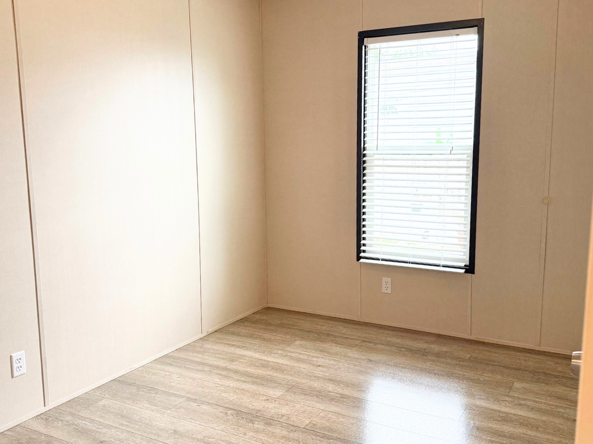 Empty room with beige walls, a single window with blinds, wood-look flooring, and two electrical outlets.